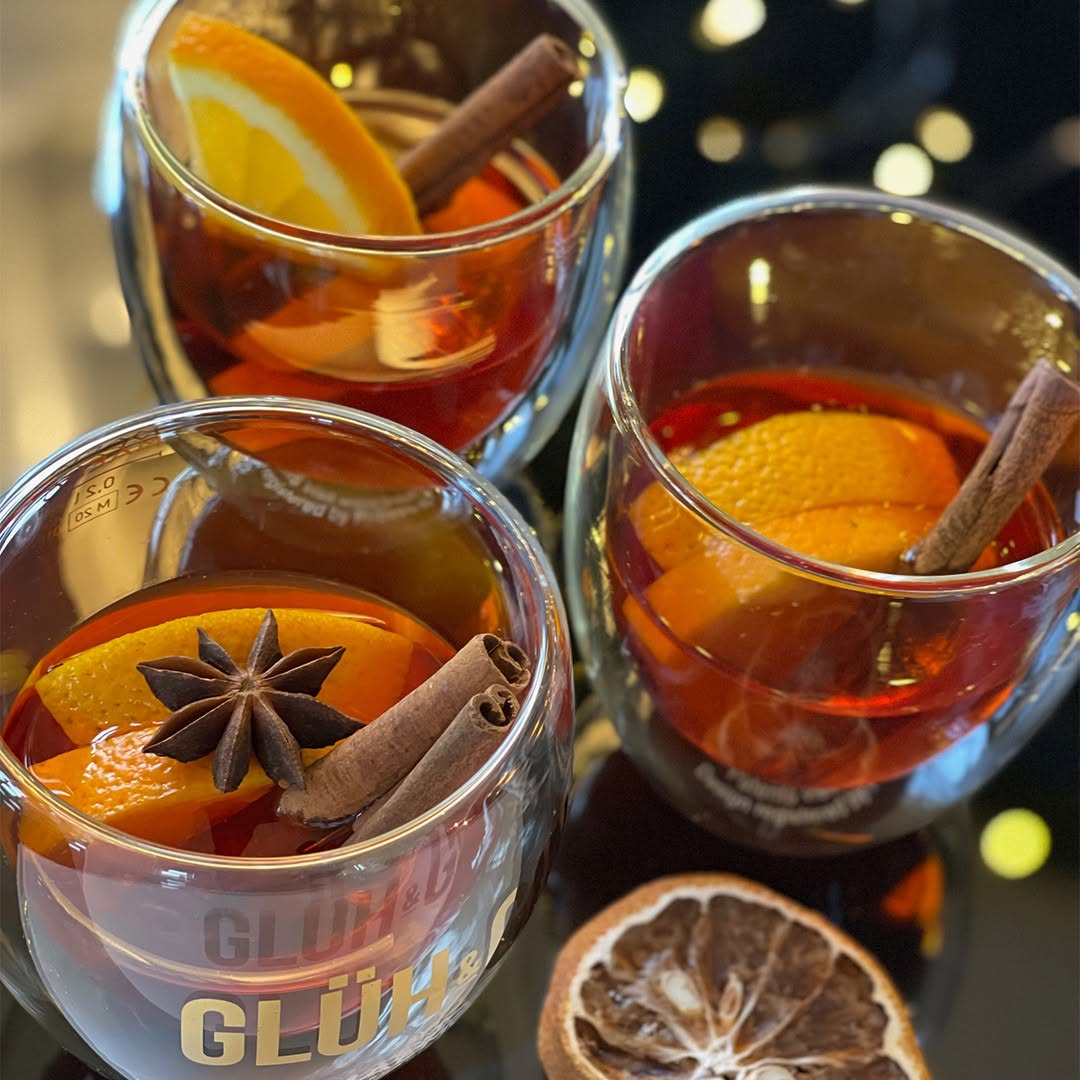 Drei Glaeser Glueh & Gin von MAEMO, gefuellt mit wuerzigem Winterdrink aus Orangenscheiben, Zimtstangen und Sternanis, stilvoll serviert vor unscharfem Lichterhintergrund – ein Genuss fuer kalte Tage.