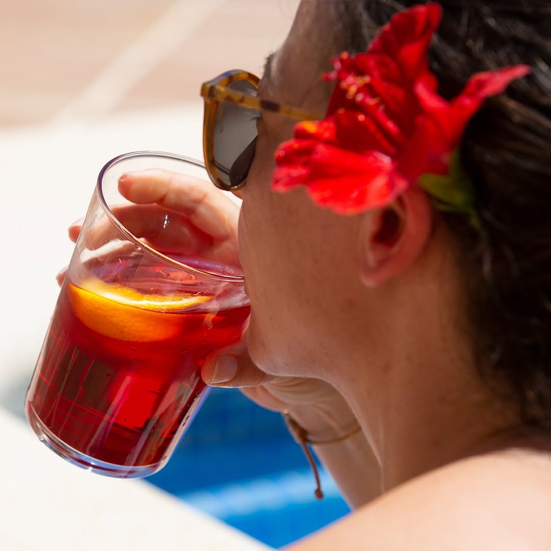 Frau mit Sonnenbrille und roter Hibiskusblume im Haar geniesst ein Glas MAEMO Berry mit Orangenscheibe am Pool – erfrischender Bio-Drink in sommerlicher Atmosphaere.