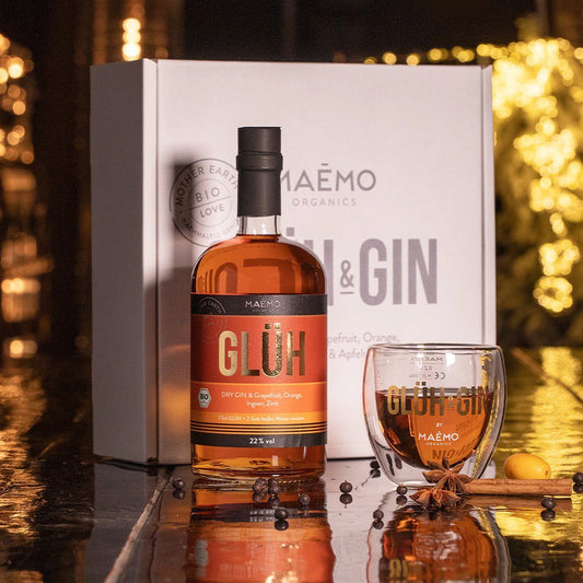 Stimmungsvolle Aufnahme der MAEMO Glueh & Gin Genießerbox mit einer Flasche Bio Dry Gin (22 % vol) sowie einem doppelwandigen Glas. Dekoriert mit Wacholderbeeren, Zimtstange, Sternanis und Zitrusfrucht vor festlichem Hintergrund – ideal als Geschenk oder fuer gemuetliche Winterabende.
