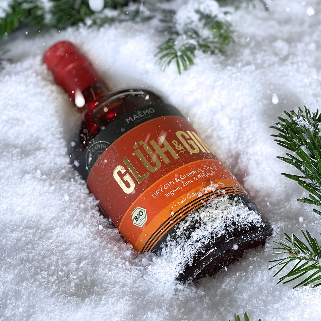 Flasche MAEMO Glueh & Gin (Bio Dry Gin mit Grapefruit, Orange, Ingwer, Zimt und Apfelsaft) liegt im frischen Schnee, dekoriert mit Tannenzweigen und fallenden Schneeflocken – perfekte Winterstimmung.