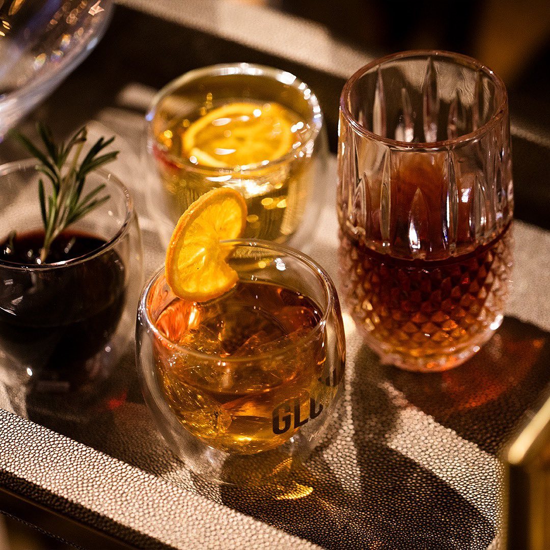 Vier elegante Cocktailglaeser mit verschiedenen Glueh & Gin Variationen auf einem Tablett: goldene und rubinrote Drinks mit Garnituren wie Orangenscheibe und Rosmarinzweig, stilvoll inszeniert in warmem Licht.